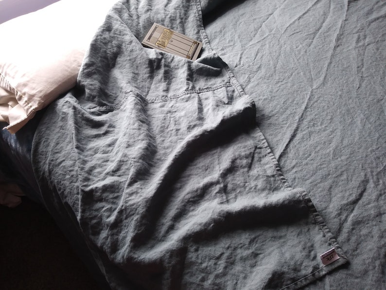 May include: A crumpled, slate-blue linen sheet covers a bed, with a pillow visible in the upper left corner. A small tag is visible on the sheet. A folded section of the sheet has a pocket with a small card inside.