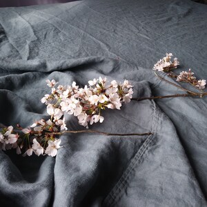 May include: A close-up of a blue-grey linen fabric with a branch of white cherry blossoms. The fabric is draped, creating soft folds and shadows. The blossoms are in full bloom.