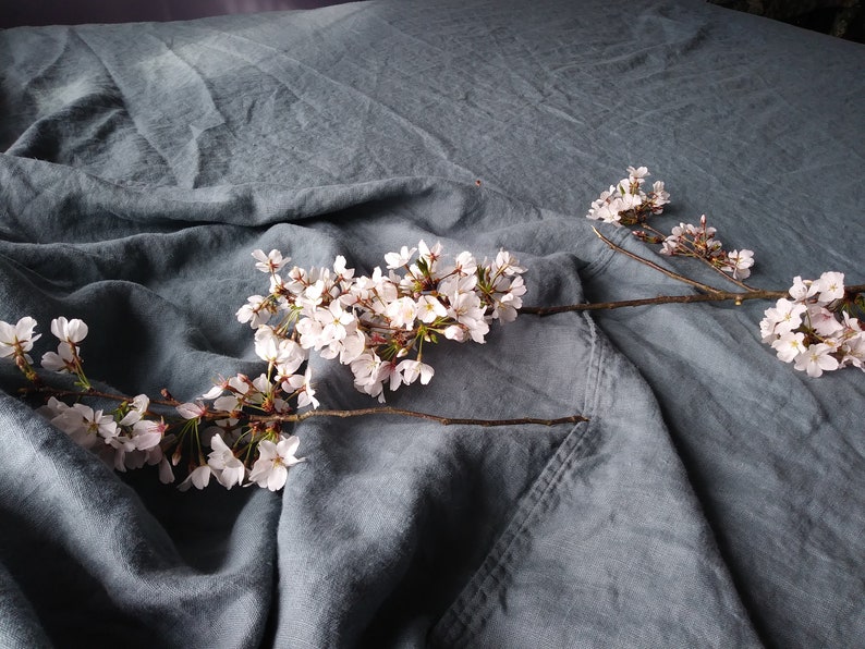 May include: A close-up of a blue-grey linen fabric with a branch of white cherry blossoms. The fabric is draped, creating soft folds and shadows. The blossoms are in full bloom.