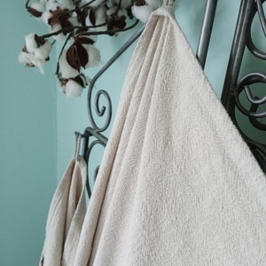 May include: Beige textured towels hanging on a decorative metal rack. The towels have a woven pattern and are looped over the rack. Cotton branches with white blooms are in the background, against a light blue wall.