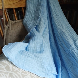 May include: A light blue, crinkled fabric blanket draped over a wooden chair. The blanket is made of a soft material and has a textured appearance. A beige tote bag hangs on the chair. The chair is in front of a bookshelf.