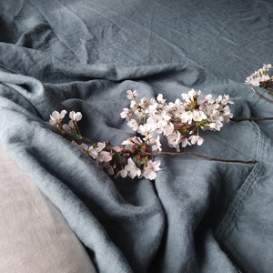 May include: A close-up of a blue-green linen fabric with a soft, wrinkled texture. A branch of cherry blossoms with delicate white flowers and brown stems rests on the fabric, creating a serene and natural composition.