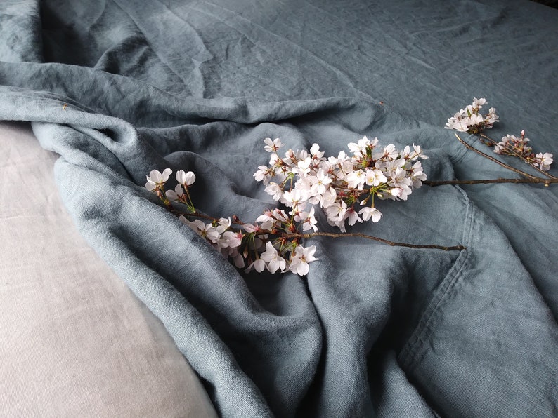 May include: A close-up of a blue-green linen fabric with a soft, wrinkled texture. A branch of cherry blossoms with delicate white flowers and brown stems rests on the fabric, creating a serene and natural composition.