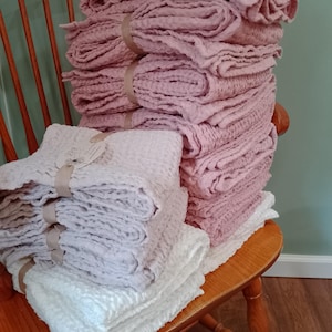 May include: Stacks of folded, textured towels in shades of pink, white, and lavender are bundled with ribbon and arranged on a wooden chair. The towels have a waffle-weave pattern, suggesting a spa or home decor theme.