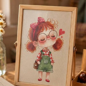 May include: A cross-stitch embroidery in a light wood frame depicting a cartoon character with red pigtails, round glasses, a red plaid shirt, and green overalls. The character has a red bow and a small red heart floating nearby. Text reads Patterns by Viktoriya Ivchenko.