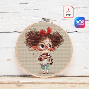 Puede incluir: Patrón de punto de cruz que presenta a una niña de dibujos animados con cabello castaño rizado, gafas y un lazo rojo. La niña lleva un suéter a rayas, pantalones azules y zapatos rojos. El diseño está en un bastidor de bordado de madera.