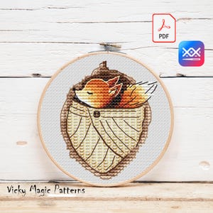 May include: Cross stitch pattern featuring a sleeping fox curled up inside a brown leaf. The fox is orange and white with a brown nose and black eyes. The pattern is in a round embroidery hoop.