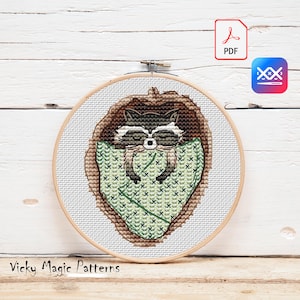 May include: A cross-stitch pattern featuring a raccoon sleeping in a green and brown nest. The pattern is in a round embroidery hoop and is available as a PDF download.