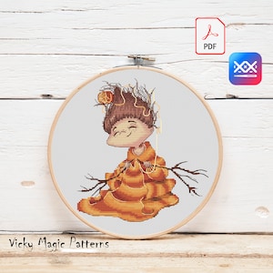 May include: Cross stitch pattern of a smiling character with brown hair, wearing an orange dress and holding a ball of yarn. The character is surrounded by branches and leaves. The pattern is in a white embroidery hoop.