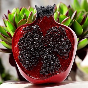 May include: A glass pomegranate sculpture, cut in half, showing dark, round seeds. The exterior is a deep red with a black stem. The background features green succulent plants, creating a contrast of colours and textures.