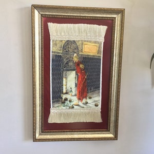 May include: A framed tapestry with a gold frame and a red mat. The tapestry depicts a person in a red robe standing in a doorway with a basket in their hand. The tapestry has a white fringe border.