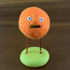 May include: A whimsical orange fuzzy character with blue eyes and a black mouth, mounted on thin copper wire legs. The legs are attached to a small, rounded, light green base. The figure has a surprised expression.