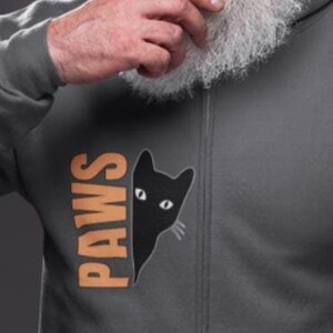 May include: A grey zip-up hoodie with a black cat peeking out from behind the word "PAWS" printed in orange on the left side of the chest.