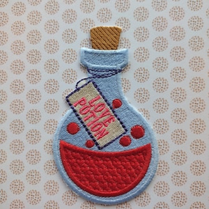 May include: A blue glass bottle with a cork stopper and a red liquid inside. The bottle has a tag that says "Love Potion" on it.