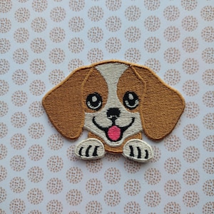 May include: Embroidered patch of a brown and white beagle puppy with black eyes and a pink tongue sticking out. The puppy is looking directly at the viewer with its paws in the air.