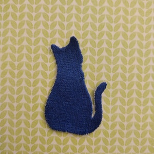 May include: A blue felt cat silhouette, sitting with its tail curled around its legs. The cat is on a green and white patterned background.