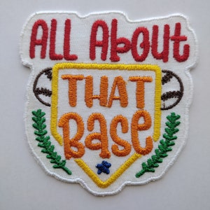 May include: White embroidered patch with a baseball diamond shape. The patch says "All About That Base" in red, orange, and yellow lettering. There are green leaves on either side of the diamond and a blue star in the center.