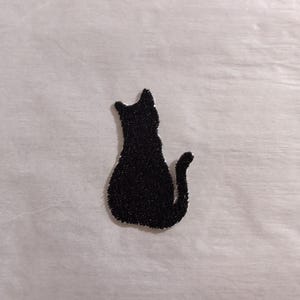 May include: A black embroidered patch in the shape of a sitting cat silhouette. The cat is depicted from behind, showing its pointed ears, rounded back, and a tail that curves slightly upward. The patch has a textured surface and defined edges.