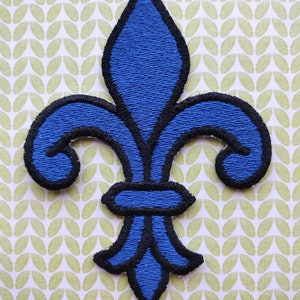 May include: A blue embroidered fleur-de-lis patch with black outlines. The patch is on a green and white patterned background.