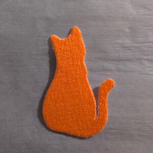 May include: A bright orange embroidered patch in the shape of a sitting cat seen from behind. The cat silhouette features pointed ears and a curved tail extending to the right. The fabric texture of the embroidery is clearly visible throughout.