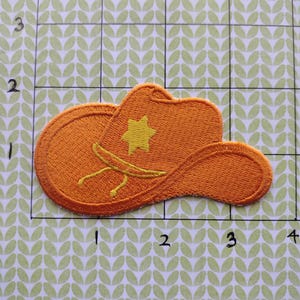 May include: An embroidered patch in the shape of a cowboy hat. The hat is orange with a yellow star and a yellow tie. The patch is on a green and white patterned background.