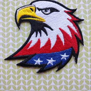 May include: Embroidered iron-on patch of a bald eagle with red, white, and blue colors. The eagle's head is facing left and has a yellow beak. The eagle's wing is in the shape of the American flag.