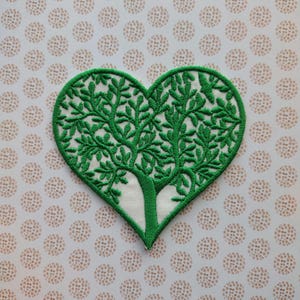 May include: Green embroidered heart-shaped patch with a tree design. The tree has a white trunk and green leaves.