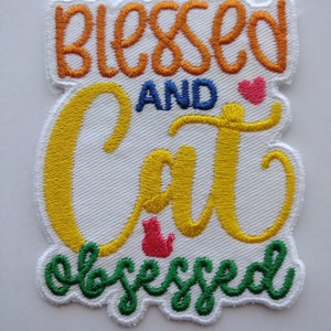 May include: A white embroidered patch with the text "Blessed and Cat Obsessed" in yellow, orange, green, and blue thread. A small red heart and a red cat silhouette are also embroidered on the patch.