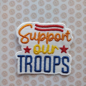 May include: White embroidered patch with red, yellow, and blue thread. The patch says "Support our Troops" with two red stars.