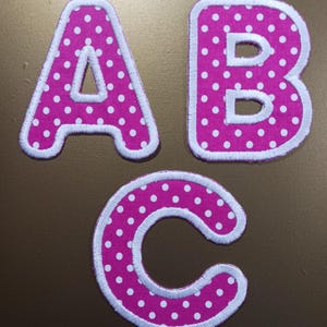 May include: Three pink fabric letters with white polka dots and white outlines. The letters are A, B, and C.