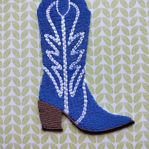 May include: Blue embroidered cowboy boot patch with white stitching. The boot has a brown heel and a pointed toe.