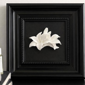 Può includere: Una cornice quadrata nera con un bordo interno perlato contenente una scultura tridimensionale di un fiore di giglio bianco centrata su uno sfondo nero solido. La cornice ha una finitura opaca liscia e un profilo profondo che mette in risalto i petali.