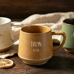 May include: Three ceramic mugs with speckled finishes in cream, tan, and sage green. The cream mug features the name Florence and a heart. The tan mug is labelled Evelyn Est. 2026. The green mug displays the name Amelia and Est. 2026.