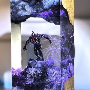 May include: A rectangular resin sculpture featuring a black figure with a red chest, standing on a dark, textured base. The sculpture is encased in clear resin with swirling purple and gray accents, and gold-colored wood on the sides.