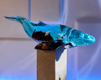 Może przedstawiać: Niebieska szklana rzeźba wieloryba z ciemną podstawą, zamontowana na kwadratowym, jasnym cokole. Kształt wieloryba jest opływowy, z zakrzywionym ciałem i uniesionym ogonem. Rzeźba jest eksponowana na białym i niebieskim tle.