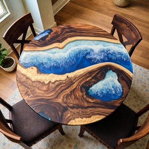Puede incluir: Mesa de comedor redonda de madera con un diseño único de resina epoxi. La mesa presenta un patrón de olas azules y blancas en el centro, rodeado de una rica veta de madera marrón. Cuatro sillas de madera con asientos marrones oscuros rodean la mesa.