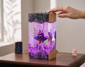Könnte beinhalten: Eine rechteckige Harzlampe mit dunkler, strukturierter Basis und Oberseite sowie einem klaren Mittelteil. Im Inneren des Harzes befindet sich eine schwarz-violette Superheldenfigur auf einer Felsformation, beleuchtet durch ein violettes Licht.