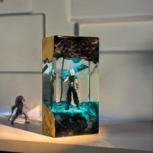 May include: A rectangular resin art piece featuring a figurine of a character with a large sword. The resin is clear with blue and black accents, and the base has a gold and brown textured finish. A second figurine is in the background.
