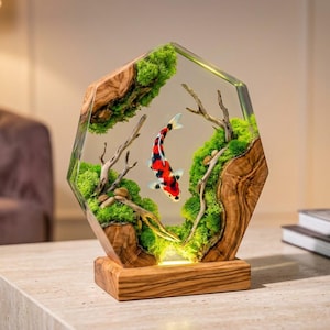 May include: A hexagonal resin and wood sculpture featuring a central koi fish with red, white, and black markings. The piece incorporates natural wood grain, bright green moss, and small driftwood branches encased in clear resin on a wood base.