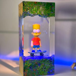 May include: A clear resin block featuring a Bart Simpson figurine on a skateboard. The figure is wearing a red shirt and blue shorts. The base of the block has green and red foliage, with a blue light illuminating the scene. The top of the block has green foliage.