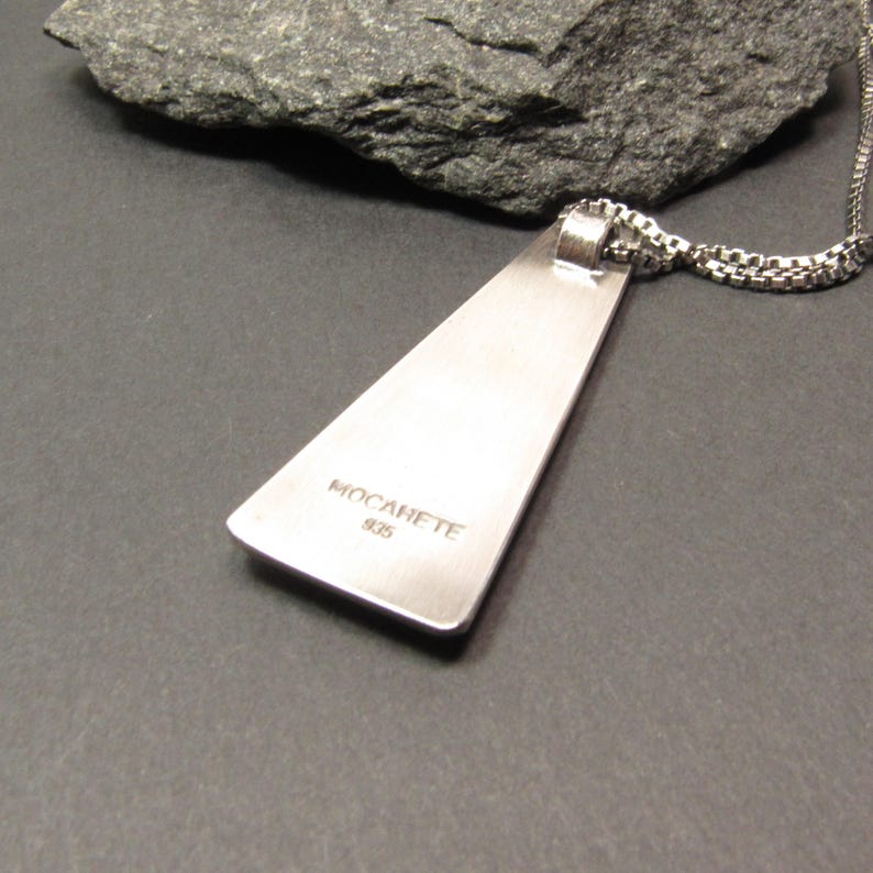 Puede incluir: Collar con colgante de plata con un colgante triangular. El colgante tiene las palabras "MOCARETE 925" grabadas en la parte posterior.