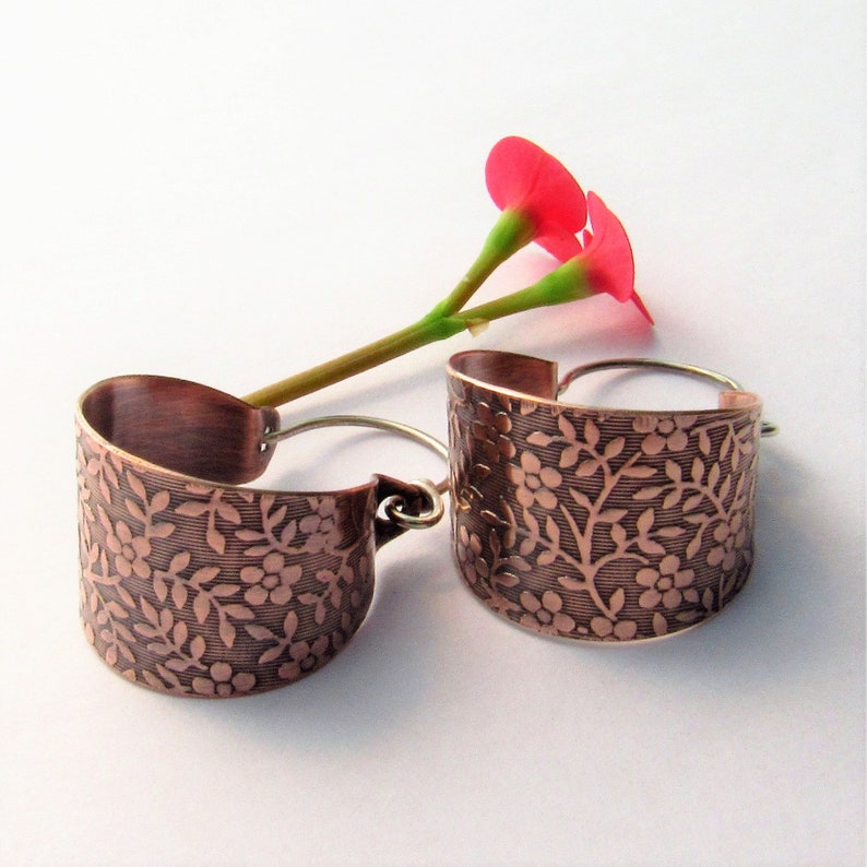 May include: A pair of copper hoop earrings with a floral design. The earrings are a light copper color and have a hammered finish. The floral design is embossed into the metal and features small flowers and leaves.