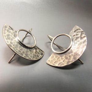May include: A pair of silver earrings with a hammered finish and a circular cutout. The earrings are shaped like a crescent moon.