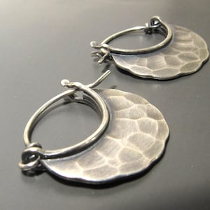May include: A pair of silver-toned hoop earrings. Each earring features a hammered, crescent-shaped charm attached to a circular hoop. The metal has a textured surface and a slightly darkened finish, creating a vintage aesthetic. The earrings are displayed against a dark background.