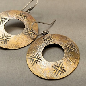 May include: A pair of handcrafted, bronze-colored earrings. Each earring is a circular disc with a central hole and embossed cross-like designs. The earrings have a textured surface and are suspended from silver-colored ear wires.