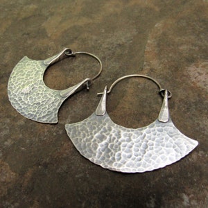 May include: A pair of silver hoop earrings with a hammered, fan-shaped design. The earrings are made of metal and have a textured finish.