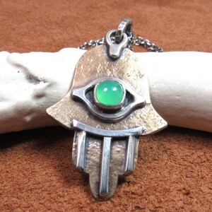 May include: A silver and bronze-toned Hamsa hand pendant necklace with a green gemstone center. The pendant has an eye design and is suspended from a silver chain. The pendant is approximately 2 inches in length.