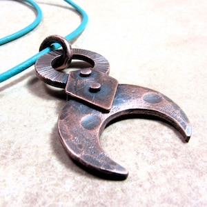 May include: A handcrafted copper pendant necklace featuring a crescent moon design. The pendant is attached to a turquoise leather cord. The copper has a textured finish and is detailed with rivets and circular accents.