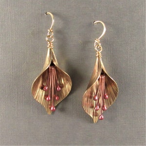 May include: A pair of gold-toned earrings with a flower design. The earrings have a delicate, layered petal design and feature small, pink beads hanging from the center.