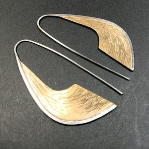 May include: A pair of modern earrings with a silver hook and a gold-colored, textured, leaf-shaped design. The earrings have a two-tone metallic finish and a minimalist aesthetic. The jewelry is displayed on a dark surface.